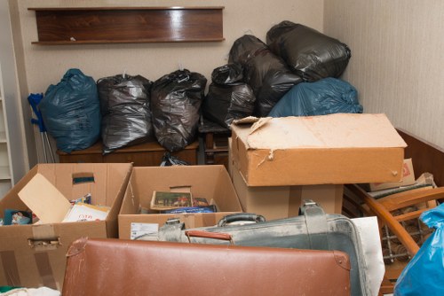 A cluttered indoor space with several large black and blue rubbish bags stacked against a wooden shelf on the wall, likely containing waste materials. In the foreground, there are open cardboard boxes filled with various paper items, possibly magazines or documents, and a metallic rubbish bin lying on its side nearby. To the right, a folded wooden chair and some flattened cardboard boxes are visible, with one box partially torn. The environment appears to be a storage or basement area with plain beige walls and a tiled floor, indicating it is used for waste collection or storage before disposal. This scene relates to the services provided by Business Waste Removal Battersea, assisting with the removal of domestic or business rubbish in the SW11 postcode district, including areas around Battersea and South West London.