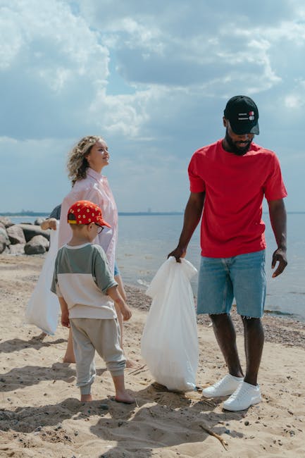 A man and two children on a sandy beach engaged in a rubbish collection activity. The man, dressed in a red t-shirt, light blue denim shorts, and a black cap, is holding a large white waste bag, appea