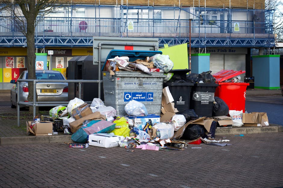 A man and two children on a sandy beach engaged in a rubbish collection activity. The man, dressed in a red t-shirt, light blue denim shorts, and a black cap, is holding a large white waste bag, appea